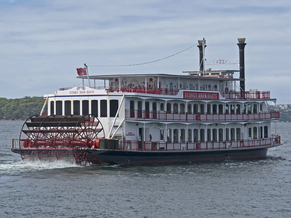 The red-and-white paddle-wheel showboat Sydney Showboat II on Sydney Harbour in daylight, with its red paddle wheel at the stern, tiers of white decks with red railings and two black funnels.